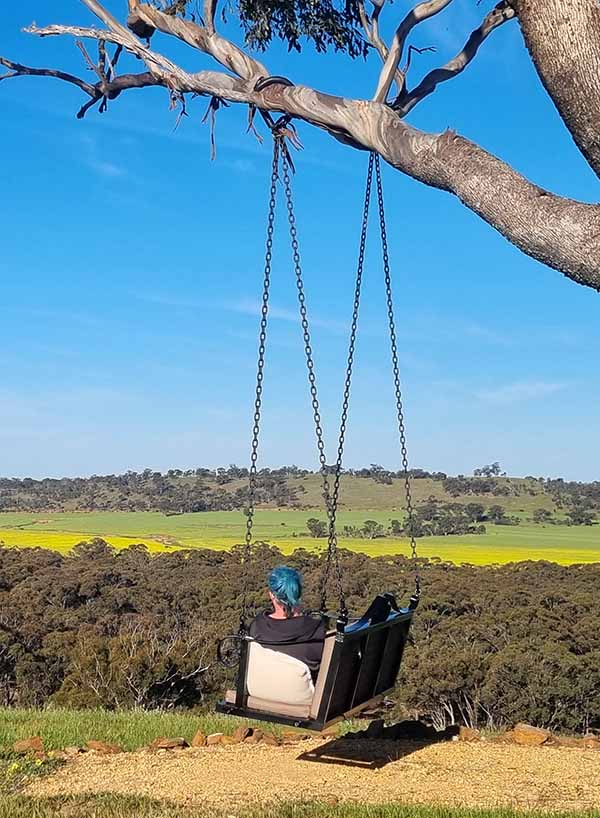A lady sitting in a swing seat, looking across a valley, that's yellow with Canola crops.